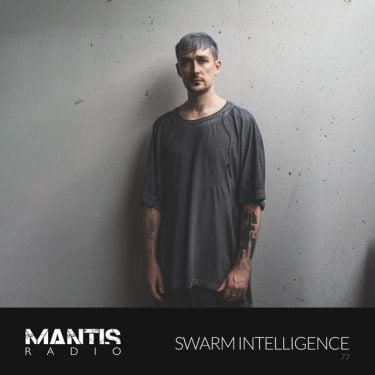 Swarm Intelligence standing in front of a wall with grey hair, and a grey long cotton vest - Mantis Radio