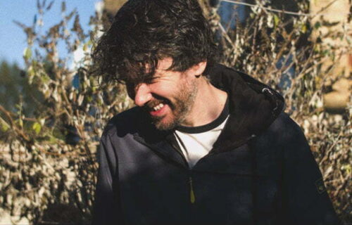 Interjector (Matt Faulkner) smiling with his head to the side in a garden with terrace housing behind him and a blue sky - Mantis Radio