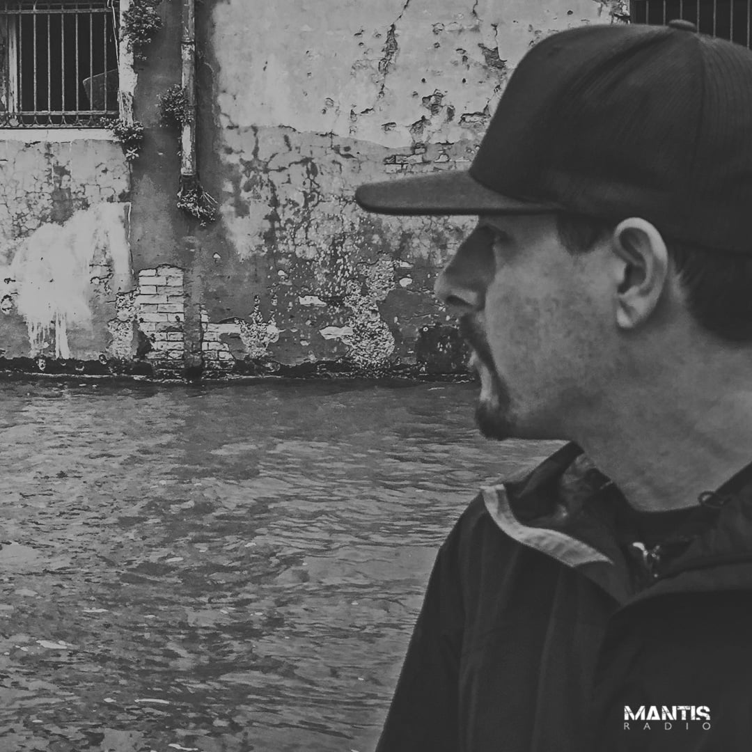 Episode guest Grym, shown in profile, wearing a baseball cap on the right side of the image, looking to the left, in front of flooded street or canal. Greyscale image.